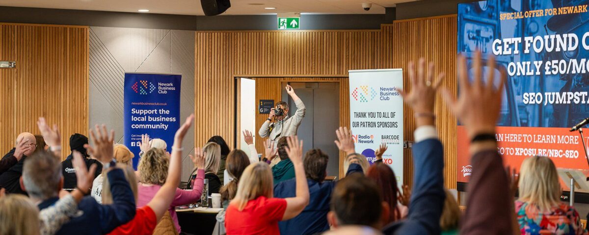 A photo taken from behind of a crowd of businesspeople at a Newark Business Club meeting at the YMCA, all with their hands raised. In the center, Nigel Lamb of Xposure Creative, holding a camera, is seen taking a photograph of the audience. Newark Business Club banners and a large presentation screen displaying "XPOSURE CREATIVE" and an "SEO JUMPSTART" offer are visible in the background.