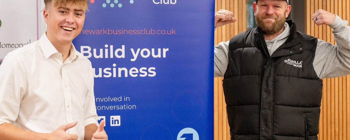 Two Newark Business Club members posing with a thumbs up and a bicep flex next to a "Build your business" banner at the YMCA.